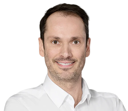 Bernhard Maier sourit à la caméra. Il porte une chemise blanche avec un col ouvert. Il a des cheveux courts brun foncé et une légère ombre de barbe.